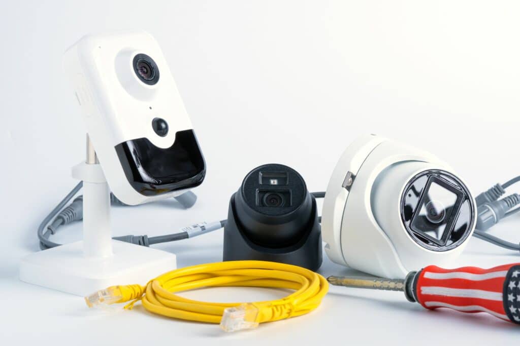 Three security cameras, one white and mounted on a stand, one black with a circular base, and one white dome camera, accompanied by a yellow Ethernet cable and a screwdriver against a white background.