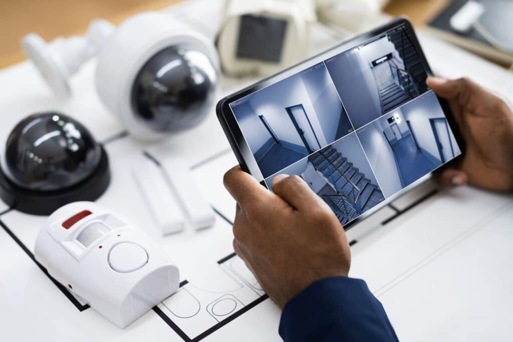 A person holding a tablet displaying security camera feeds while various security devices, including cameras and a motion sensor, are placed on a blueprint on a table.