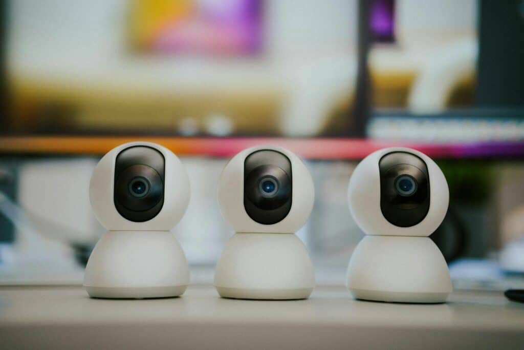 Three white security cameras with rounded designs and black lenses arranged on a desk. A blurred background shows a colorful abstract screen.