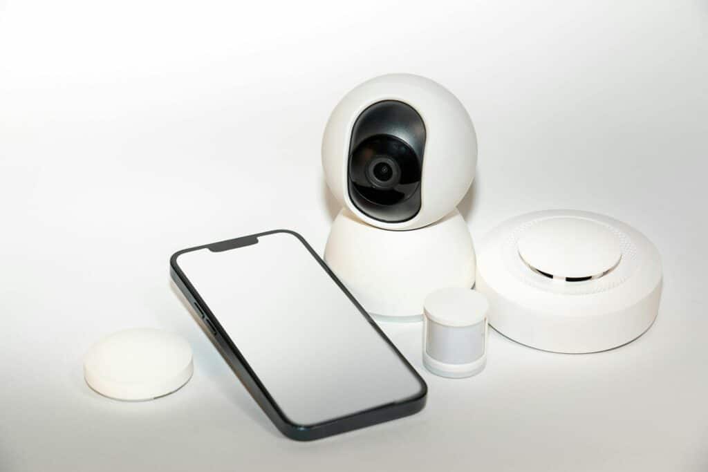 A smartphone with a reflective screen placed next to a white security camera, a motion sensor, and a smoke detector on a light background.