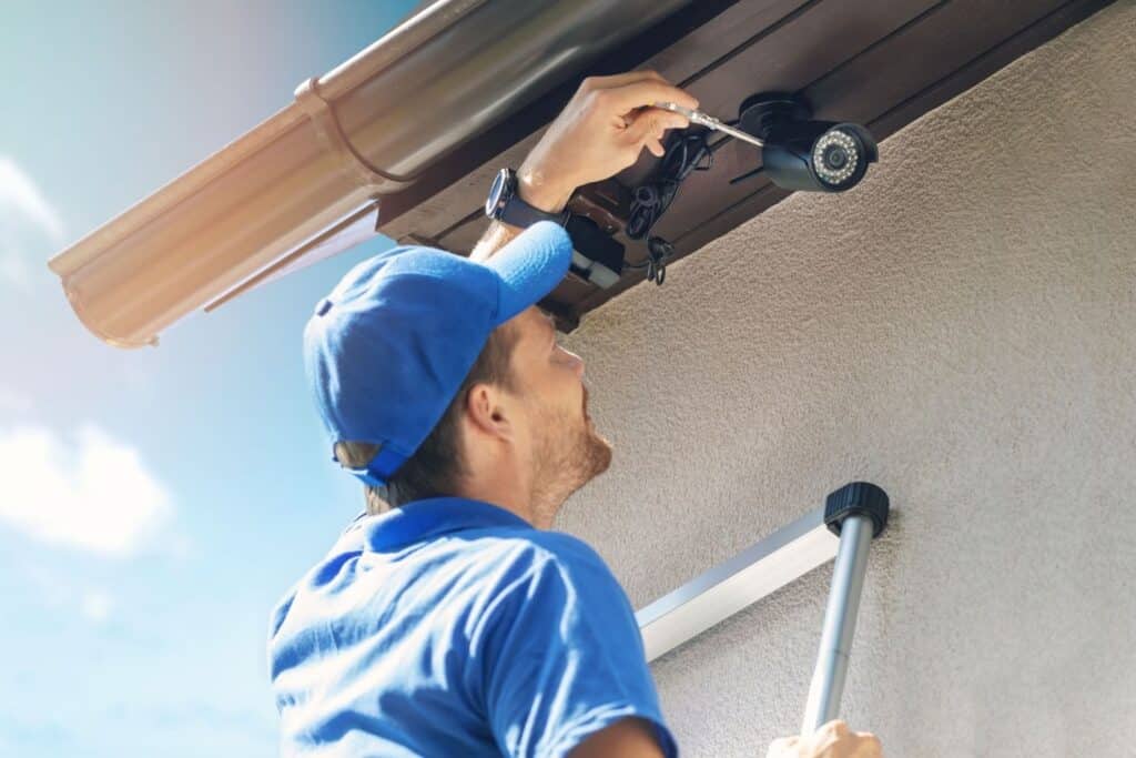 A technician wearing a blue cap and shirt installing a security camera on the eaves of a house using a ladder. The sky is clear with soft sunlight illuminating the scene.