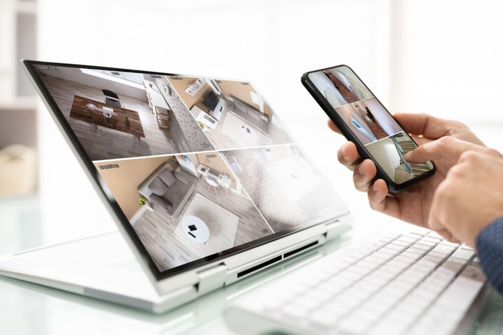 A laptop displaying multiple surveillance camera feeds beside a person holding a smartphone. A wireless keyboard is visible in front of the laptop on a glass surface.